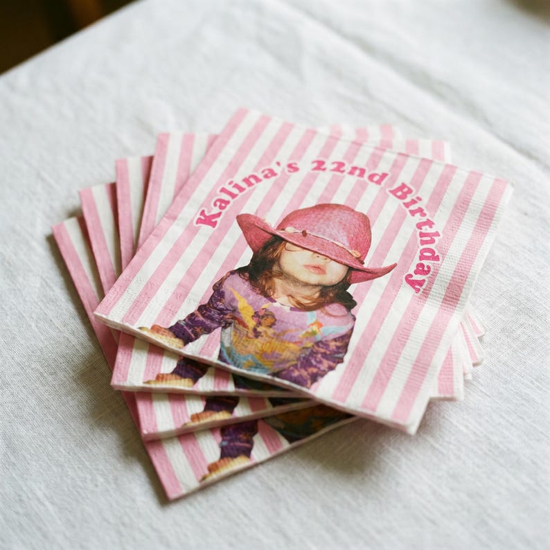 May include: A stack of square paper napkins featuring a pink and white vertical striped pattern. The center displays a graphic of a person wearing a pink cowboy hat and a patterned long-sleeve shirt, with the text Kalina's 22nd Birthday printed in pink.