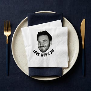 May include: A white paper napkin with a black and white image of a smiling person's face and the text "LOOK WHO'S 30!". The napkin is placed on a navy blue napkin, a speckled plate, and a gold and navy blue fork and knife.
