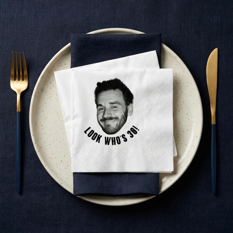 May include: A white paper napkin with a black and white image of a smiling person's face and the text "LOOK WHO'S 30!". The napkin is placed on a navy blue napkin, a speckled plate, and a gold and navy blue fork and knife.