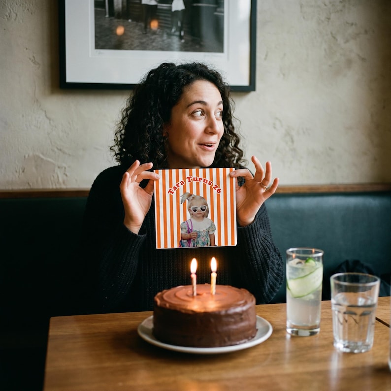 May include: A square paper napkin featuring orange and white vertical stripes with a central graphic of a child wearing sunglasses and the text Tara Turns 26. The napkin is held up behind a round chocolate birthday cake with two lit candles.