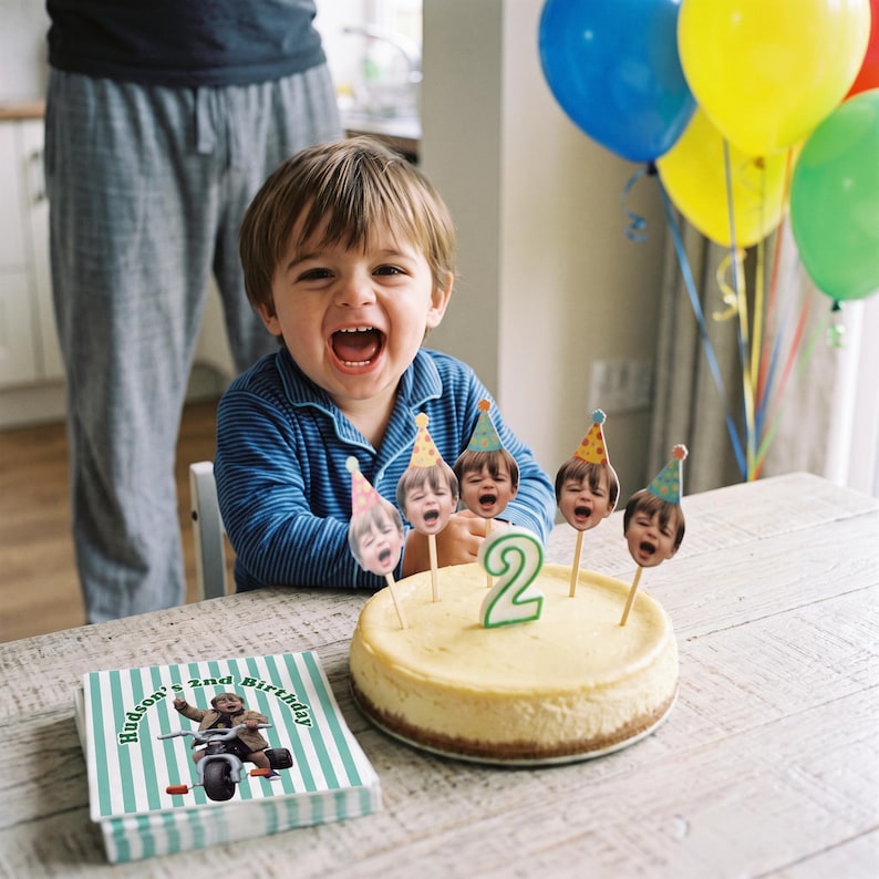 K&ouml;nnte beinhalten: Ein runder K&auml;sekuchen mit einer gr&uuml;nen Kerze in Form einer Zwei und f&uuml;nf Papier-Tortensteckern, die ein Kindergesicht mit Partyhut zeigen. Daneben liegt eine quadratische Serviette mit gr&uuml;nen Streifen und dem Text Hudson's 2nd Birthday.