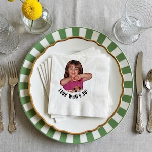 May include: A white napkin with a photo of a young girl, with the text "LOOK WHO'S 30!" on a white plate with a green and white striped border and gold trim. The plate is set with silverware and a glass.