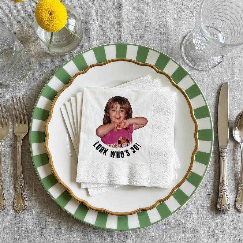 May include: A white napkin with a photo of a young girl, with the text "LOOK WHO'S 30!" on a white plate with a green and white striped border and gold trim. The plate is set with silverware and a glass.
