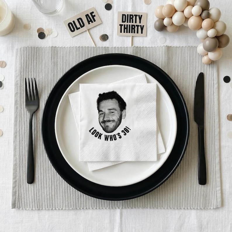 May include: A festive table setting with a black and white theme. A white napkin with a smiling face and the text "LOOK WHO'S 30!" sits on a plate. Decorative signs read "OLD AF" and "DIRTY THIRTY".