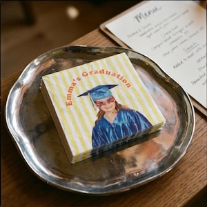 May include: A stack of square paper napkins featuring a yellow and white striped pattern. The center displays a photograph of a person in a blue graduation gown and cap, wearing star-shaped glasses, with the text Emma's Graduation printed in orange.