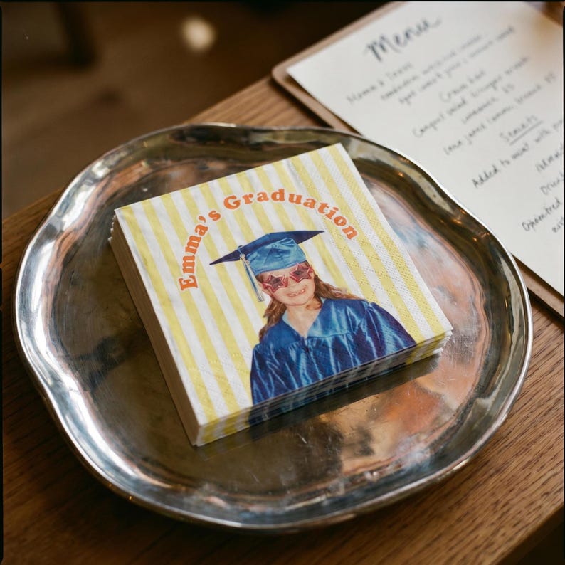 May include: A stack of square paper napkins featuring a yellow and white striped pattern. The center displays a photograph of a person in a blue graduation gown and cap, wearing star-shaped glasses, with the text Emma's Graduation printed in orange.