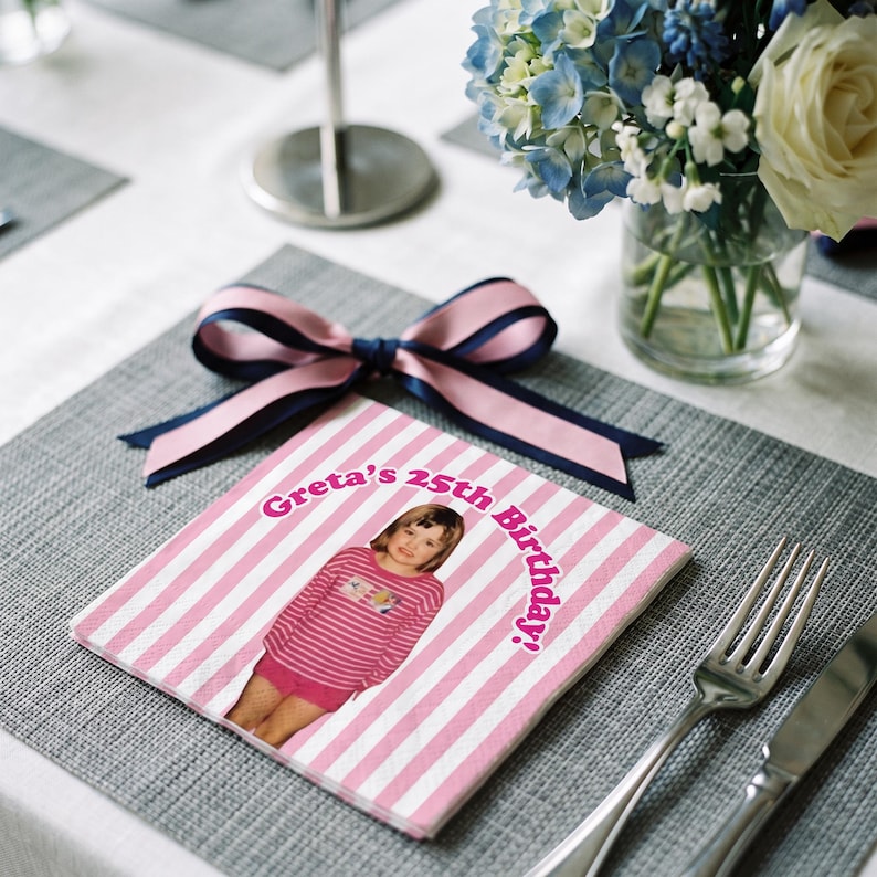May include: A square paper napkin featuring a pink and white vertical striped pattern. The center displays a photograph of a child in a pink striped shirt and shorts, with the text "Greta's 25th Birthday!" arched above. A pink and navy blue ribbon bow sits atop.