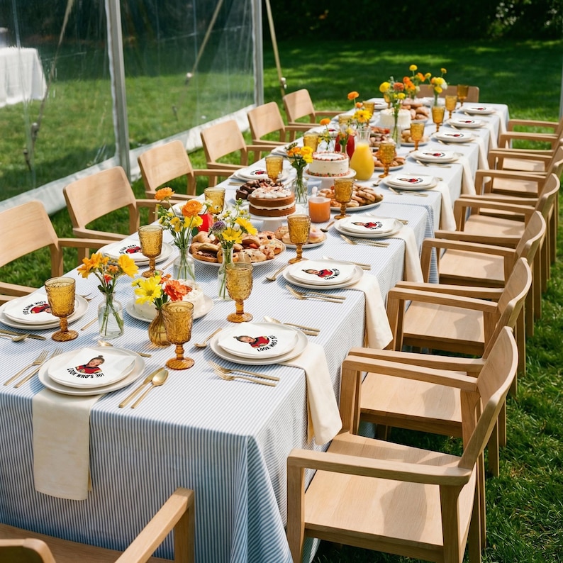 May include: A long table set for a party, covered with a blue and white striped tablecloth. The table is set with plates, glasses, and silverware. There are floral arrangements, cakes, and other food items. Wooden chairs surround the table, set on a grassy lawn.