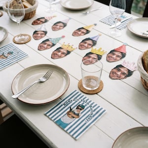 May include: A set of paper party decorations featuring cutouts of a person's face wearing various colorful party hats and crowns, alongside square paper napkins printed with blue and white stripes and the text Ruben's 30th Birthday.