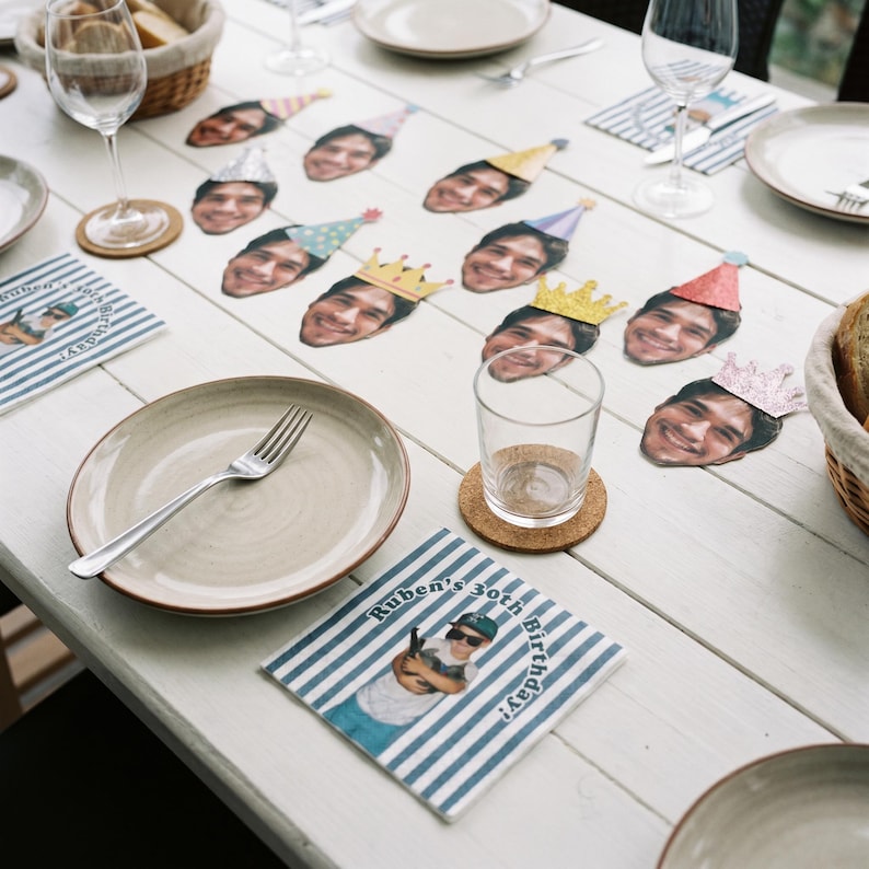 May include: A set of paper party decorations featuring cutouts of a person's face wearing various colorful party hats and crowns, alongside square paper napkins printed with blue and white stripes and the text Ruben's 30th Birthday.