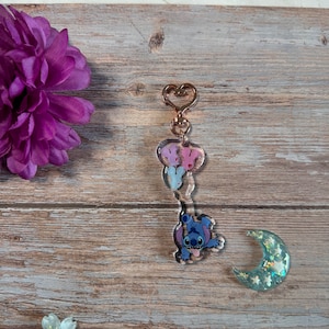 K&ouml;nnte beinhalten: Ein blauer Stitch-Charakter-Schl&uuml;sselanh&auml;nger mit einem rosa und wei&szlig;en Schmetterlingsanh&auml;nger und einem goldenen herzf&ouml;rmigen Ring. Der Schl&uuml;sselanh&auml;nger befindet sich auf einer Holzoberfl&auml;che mit einer lila Blume und einem gr&uuml;nen Mondanh&auml;nger.