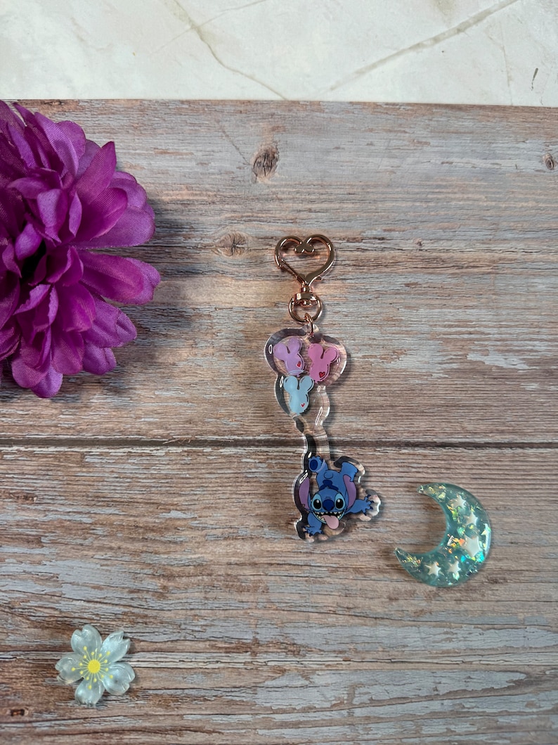 K&ouml;nnte beinhalten: Ein blauer Stitch-Charakter-Schl&uuml;sselanh&auml;nger mit einem rosa und wei&szlig;en Schmetterlingsanh&auml;nger und einem goldenen herzf&ouml;rmigen Ring. Der Schl&uuml;sselanh&auml;nger befindet sich auf einer Holzoberfl&auml;che mit einer lila Blume und einem gr&uuml;nen Mondanh&auml;nger.