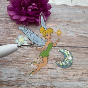 Peut inclure: Un autocollant transparent représentant Tinkerbell, une fée Disney aux cheveux blonds et une robe verte, tenant une étoile. L'autocollant a un fond de paillettes bleu et blanc et un croissant de lune avec des étoiles.