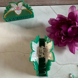 May include: Two green hair clips with white flower designs. One clip is a half-circle shape and the other is a more elongated shape.