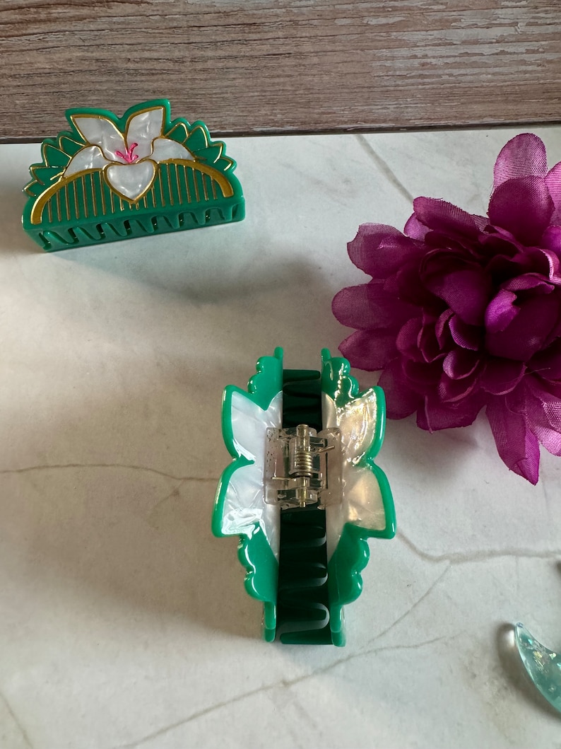 May include: Two green hair clips with white flower designs. One clip is a half-circle shape and the other is a more elongated shape.
