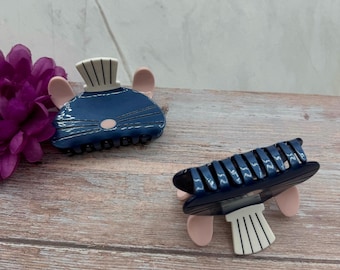 May include: Two blue and white mouse-shaped hair clips. The clips have pink ears and white stripes on the top of their heads.