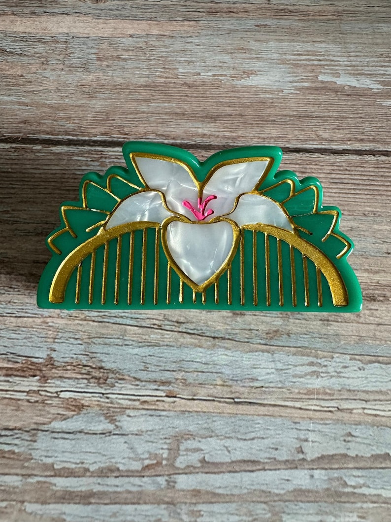 May include: A green and gold hair comb with a white flower design. The flower has a pink center.