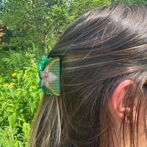 May include: A semi-circular hair claw clip in a deep green hue, featuring a central white floral motif with pearlescent petals and gold-toned linear detailing. The accessory has a scalloped edge and a structured design for securing hair.