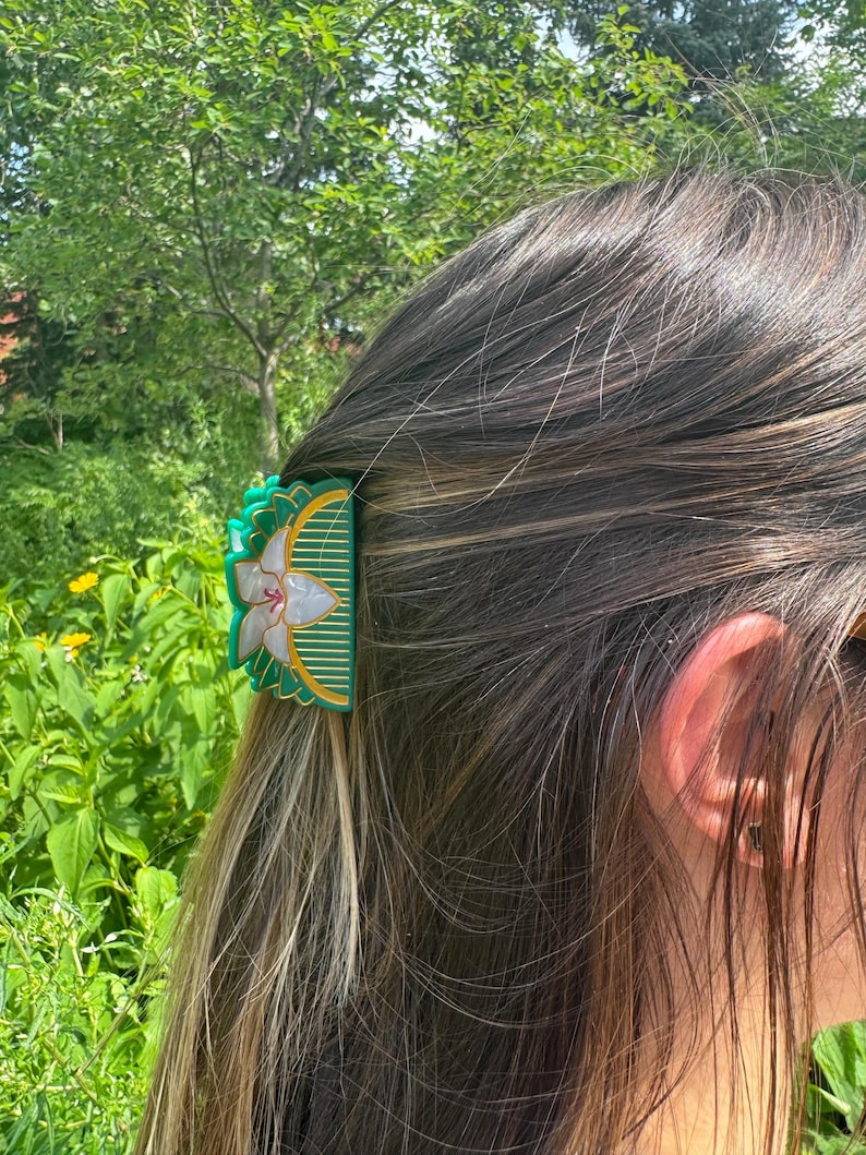 May include: A semi-circular hair claw clip in a deep green hue, featuring a central white floral motif with pearlescent petals and gold-toned linear detailing. The accessory has a scalloped edge and a structured design for securing hair.