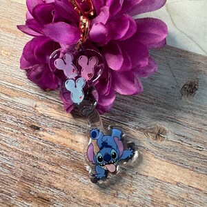 K&ouml;nnte beinhalten: Ein durchsichtiger blauer Stitch-Schl&uuml;sselanh&auml;nger mit einem rosa und wei&szlig;en Mickey-Maus-Anh&auml;nger. Der Schl&uuml;sselanh&auml;nger ist an einem goldenen Metallclip befestigt.