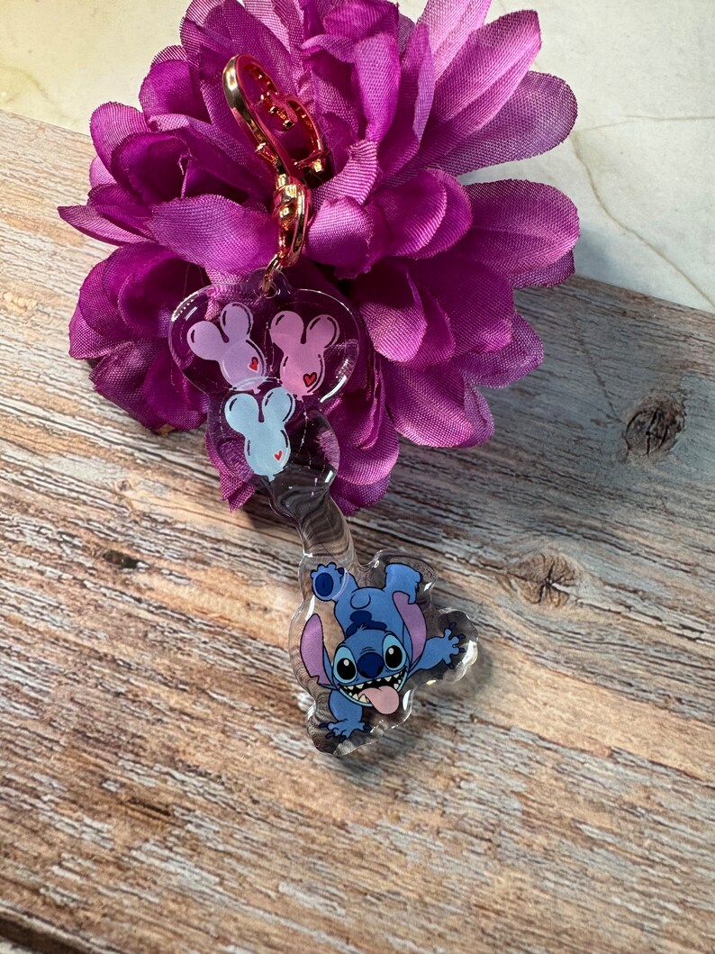 K&ouml;nnte beinhalten: Ein durchsichtiger blauer Stitch-Schl&uuml;sselanh&auml;nger mit einem rosa und wei&szlig;en Mickey-Maus-Anh&auml;nger. Der Schl&uuml;sselanh&auml;nger ist an einem goldenen Metallclip befestigt.
