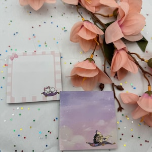 May include: Two square sticky notes with illustrations. The first note has a pink and white striped border and a white cat lying on a pink and white striped pillow. The second note has a purple background with a cartoon image of a couple riding on a magic carpet.