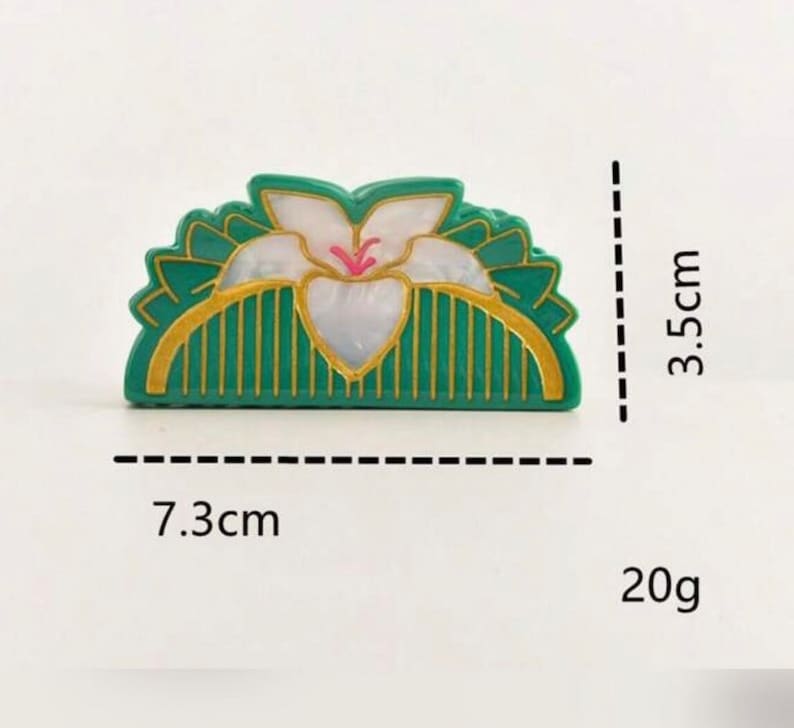 May include: A green and gold hair comb with a white flower design. The comb is 7.3 cm wide and 3.5 cm tall and weighs 20 grams.