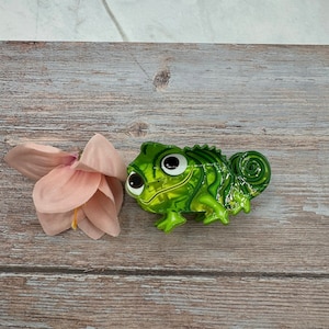 May include: A green chameleon ornament with large eyes. The ornament has a glossy finish and is next to a pink flower on a wooden surface. The chameleon's body has a textured, scale-like appearance.