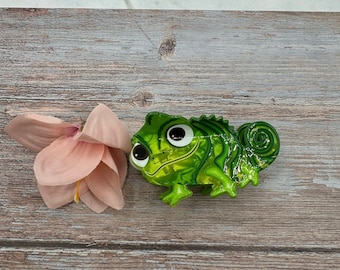 May include: A green chameleon figurine with large, expressive eyes. The figurine has a glossy finish and is positioned next to a pink flower on a wooden surface. The chameleon's body has a textured, scale-like appearance.