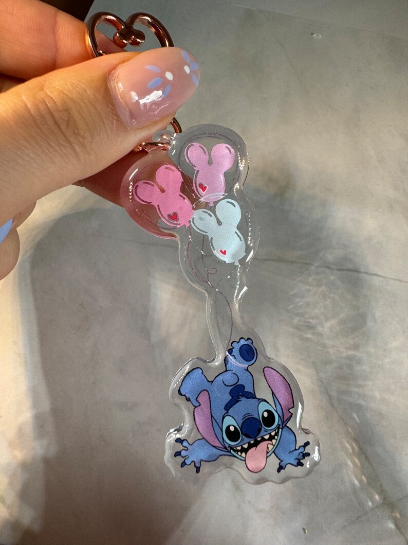 K&ouml;nnte beinhalten: Ein transparenter blauer Stitch-Schl&uuml;sselanh&auml;nger mit einem rosa, wei&szlig;en und blauen Mickey-Maus-Ballon-Anh&auml;nger. Der Schl&uuml;sselanh&auml;nger hat einen ros&eacute;goldenen herzf&ouml;rmigen Ring.