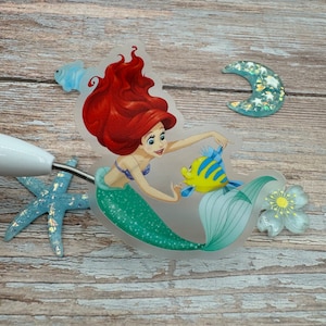 Puede incluir: Una pegatina transparente con Ariel, la Sirenita, con su cabello rojo y cola verde, nadando con un pez amarillo y azul. La pegatina est&aacute; sobre una superficie de madera.