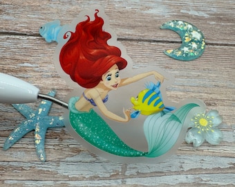Puede incluir: Una pegatina transparente con Ariel, la Sirenita, con su cabello rojo y cola verde, nadando con un pez amarillo y azul. La pegatina está sobre una superficie de madera.