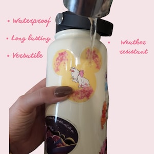 Puede incluir: Una botella de agua blanca con varias pegatinas coloridas con personajes de dibujos animados. La botella se est&aacute; enjuagando bajo un grifo. El texto "Our Stickers are: Waterproof, Long lasting, Versatile, Weather resistant" es visible en la imagen.