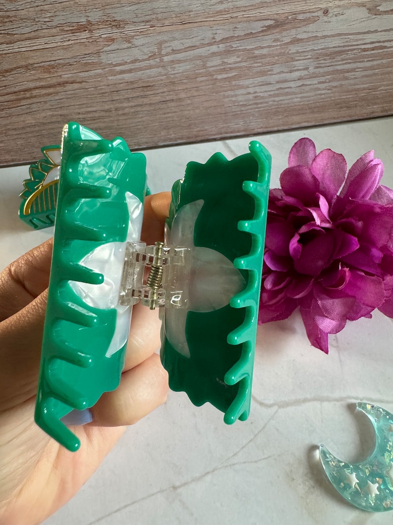 May include: A green plastic hair claw with a white swirl design. The claw is open and ready to be used to hold hair in place.