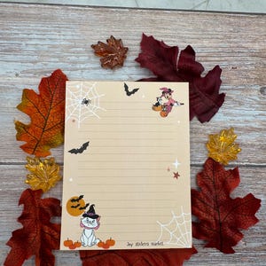 May include: A rectangular notepad with horizontal lines, featuring a white spider web, a black bat, a cartoon character in a witch hat, a white cat in a witch hat, orange pumpkins, and stars. The text "Joy stickers market" is printed at the bottom.