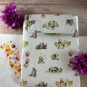 Op de afbeelding: Een witte stoffen tas met een klep sluiting. De tas is versierd met een herhalend patroon van Winnie de Poeh en zijn vrienden. De tas is perfect om kleine spullen in op te bergen, zoals sleutels, telefoon of make-up.