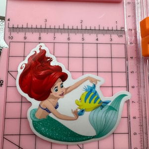 Puede incluir: Una pegatina con Ariel, la Sirenita, con su cabello rojo y su cola verde. Ella est&aacute; sosteniendo un pez amarillo con rayas azules.