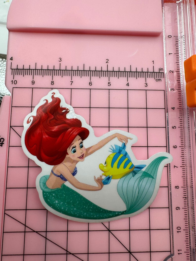 Puede incluir: Una pegatina con Ariel, la Sirenita, con su cabello rojo y su cola verde. Ella est&aacute; sosteniendo un pez amarillo con rayas azules.