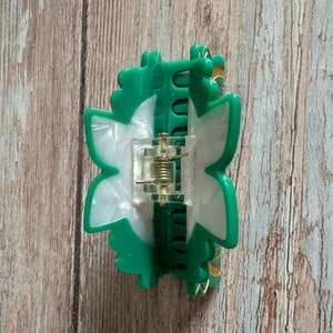 May include: A green and white plastic hair clip shaped like a flower. The clip has a spring mechanism and a gold-colored accent.