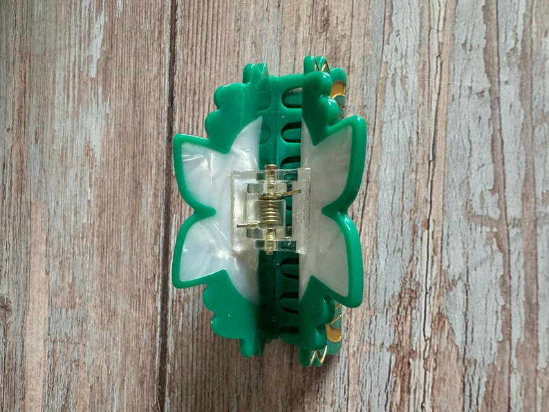 May include: A green and white plastic hair clip shaped like a flower. The clip has a spring mechanism and a gold-colored accent.