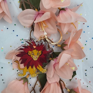 May include: A clear acrylic keychain featuring a cartoon image of Simba from Disney's The Lion King. Simba is depicted with a red mane and a yellow body, standing on a green base. The keychain has a gold metal ring.