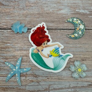 Puede incluir: Una pegatina con Ariel, la Sirenita, con su cabello rojo y su cola verde, nadando con un pez amarillo. La pegatina est&aacute; sobre un fondo de madera con otros elementos decorativos.