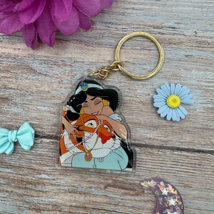 Puede incluir: Un llavero de acrílico transparente con una imagen de dibujos animados de la princesa Jasmine de Aladdin sosteniendo a Rajah el tigre. El llavero tiene un anillo de metal dorado.