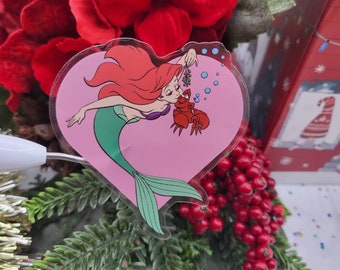 Puede incluir: Una pegatina transparente en forma de corazón con Ariel de La Sirenita. Ariel está representada con cabello rojo, una cola verde y un sujetador de concha rosa. Está besando a un cangrejo rojo con un tallo verde en su garra. La pegatina está sobre un fondo rosa con burbujas azules.
