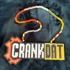 May include: A handmade beaded necklace with the text "CRANK DAT" in white block letters on a black and yellow background. The necklace has a beaded chain in shades of orange, yellow, and white. The word "CRANK DAT" is a reference to a popular dance.