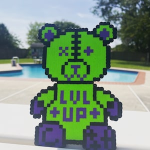 May include: Green pixelated bear with purple accents and the text "LVL UP" on the chest. The bear is made of plastic or felt and is a handmade craft.