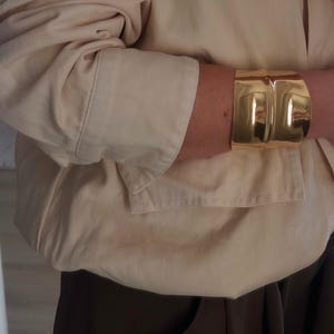 May include: A broad, gold cuff bracelet is worn on an arm. The bracelet has a smooth, reflective surface. The person is wearing a beige button-down shirt and dark brown trousers.