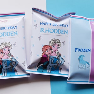 Peut inclure: Trois sachets de chips blancs et bleus sur le thème de La Reine des Neiges. Le devant des sachets représente Elsa et Anna du film La Reine des Neiges. Les sachets portent l'inscription "Happy Birthday Rhodden". Le dos d'un sachet est rose avec l'inscription "Frozen" et une image d'un cheval. Le sachet comporte également un code-barres et l'inscription "Ingrédients".