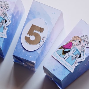 Peut inclure: Trois boîtes de fête bleues et violettes sur le thème de La Reine des Neiges. Les boîtes présentent des images d'Elsa et d'Anna du film La Reine des Neiges. Une boîte porte le numéro 5 en or. Les boîtes portent l'inscription "Tehiilah-Meyer".
