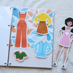 Puede incluir: Una muñeca de papel con cabello castaño y un vestido rosa, junto con una variedad de opciones de ropa que incluyen una camisa verde, una camisa azul, una camisa amarilla, una falda marrón y un mono naranja. Las piezas de ropa están sobre un fondo blanco con un patrón de remolino azul y naranja.